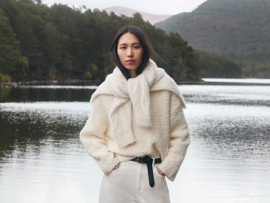 a woman in a white sweater and pants standing by a lake