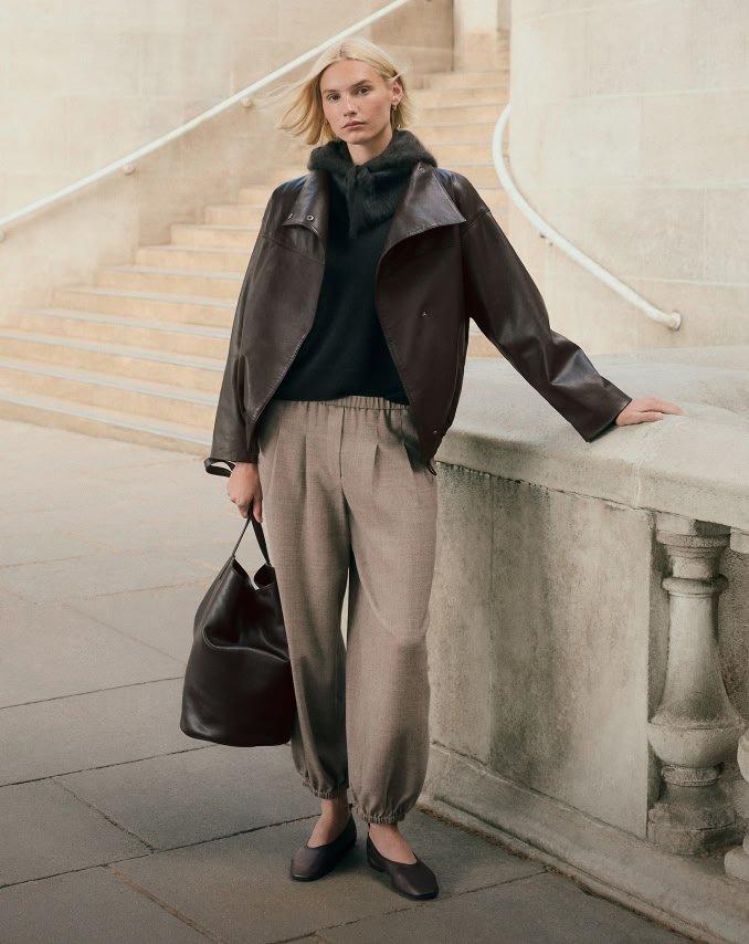 a woman in a jacket and pants leaning against a wall