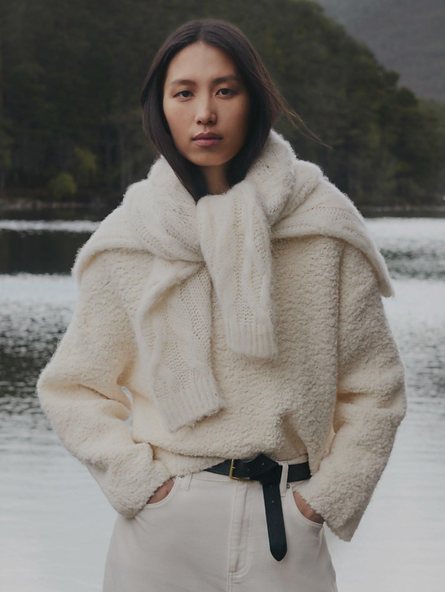 a woman in a white sweater and pants standing by a lake